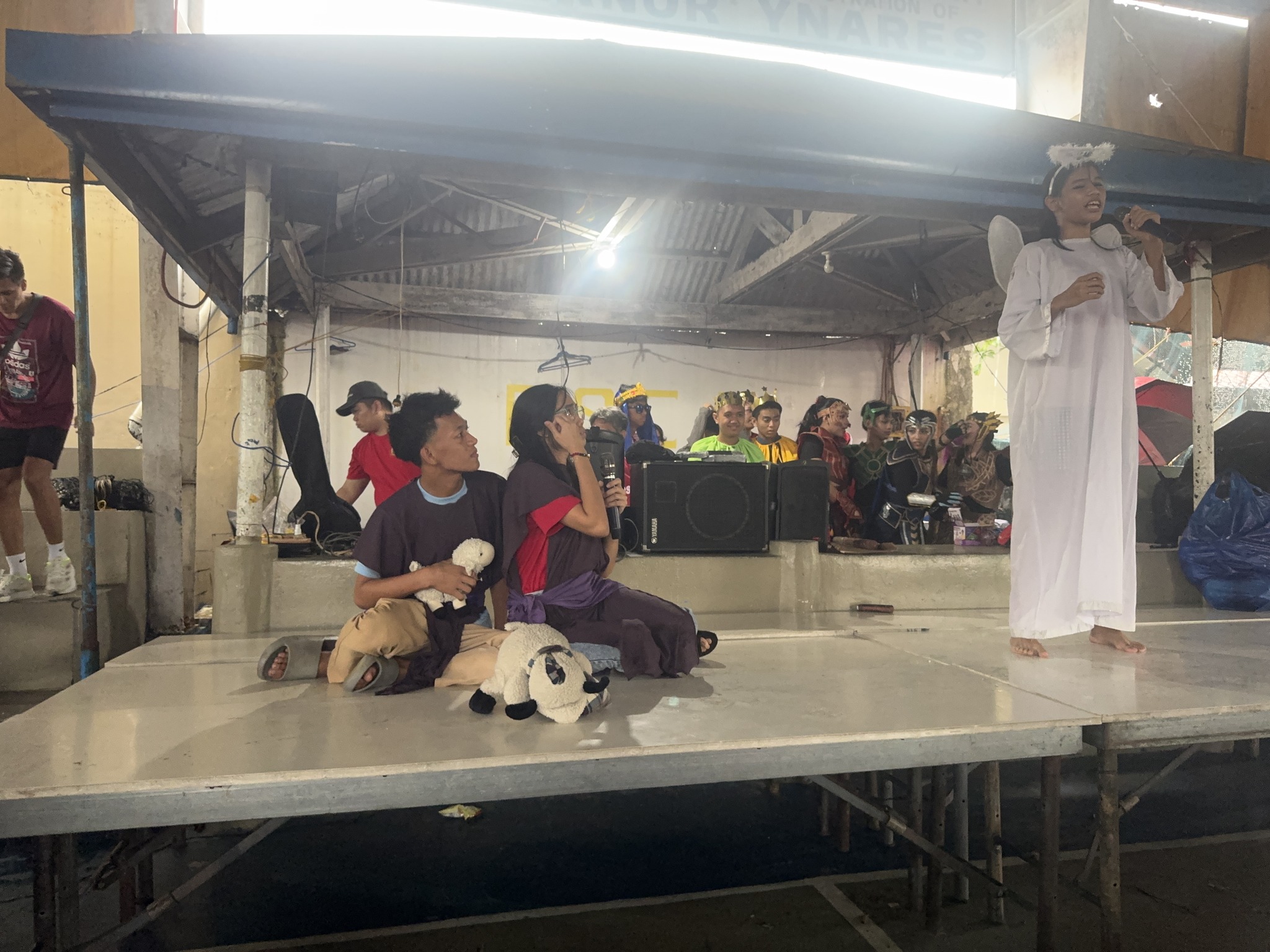 Performers on stage acting out the nativity scene with shepherds, sheep props, and an angel in white