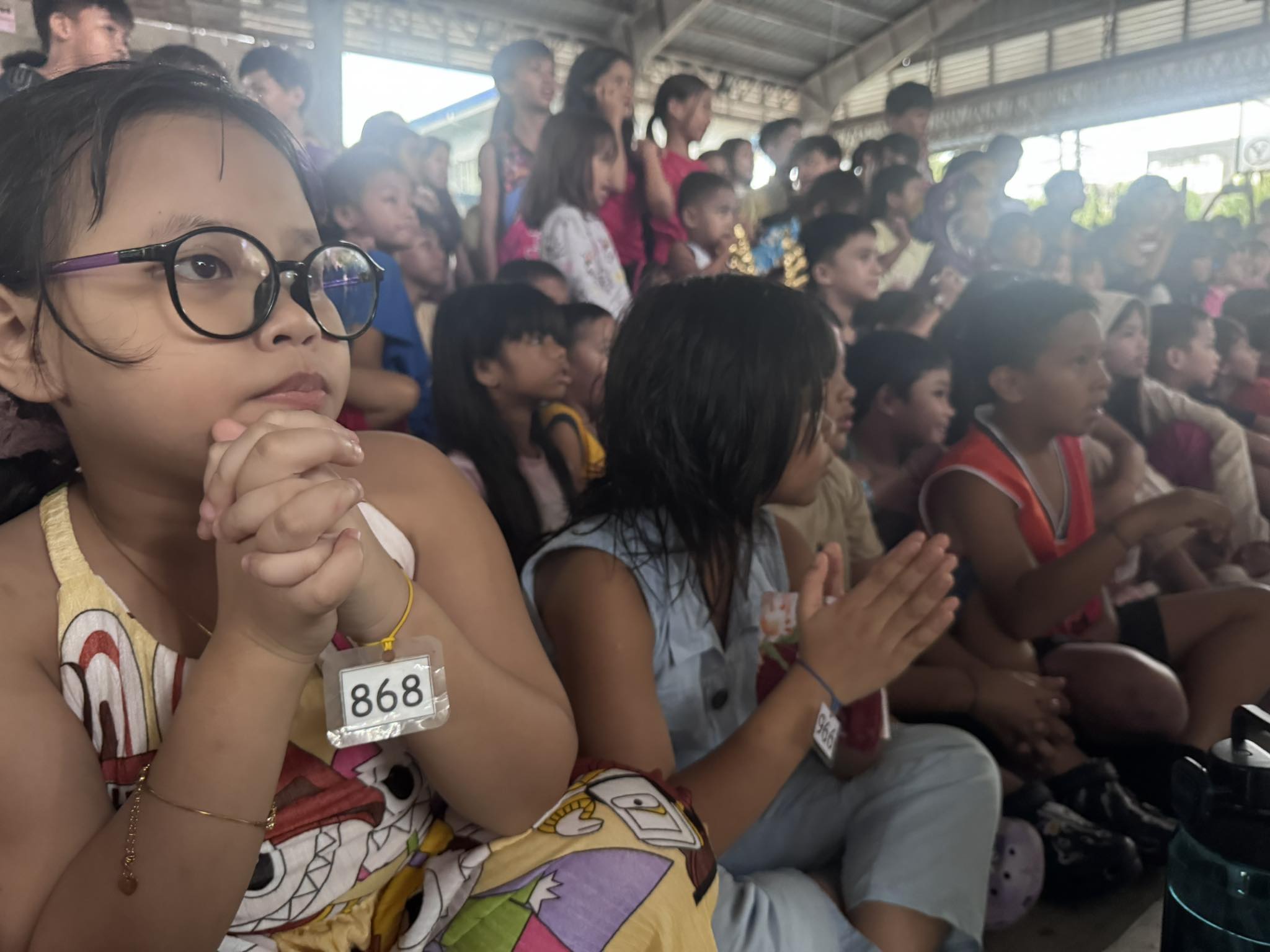 A young girl with glasses and number tag 868 sitting with hands clasped, watching the program